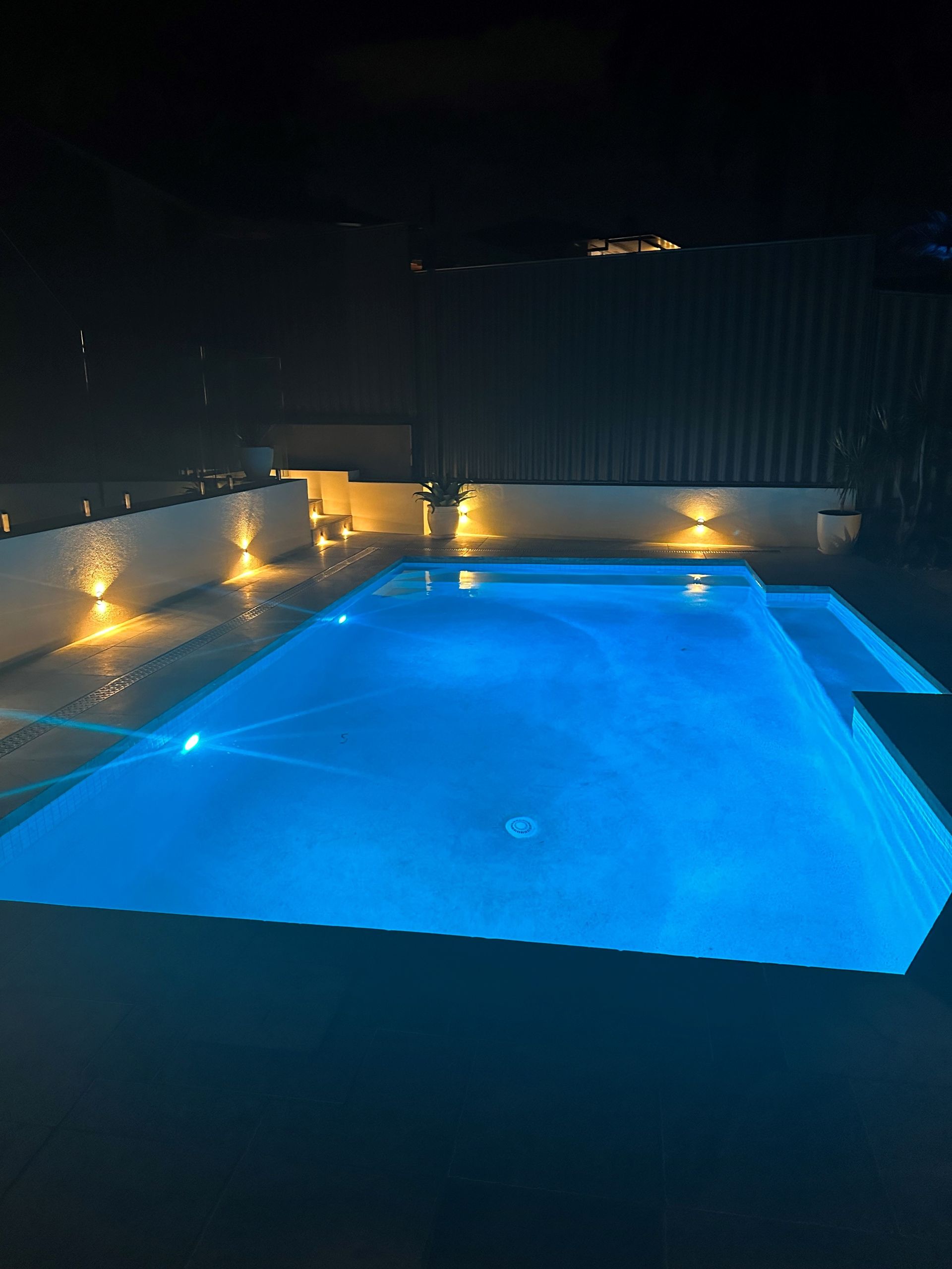 Nighttime view of a blue-lit swimming pool with surrounding illuminated white walls, backyard setting.