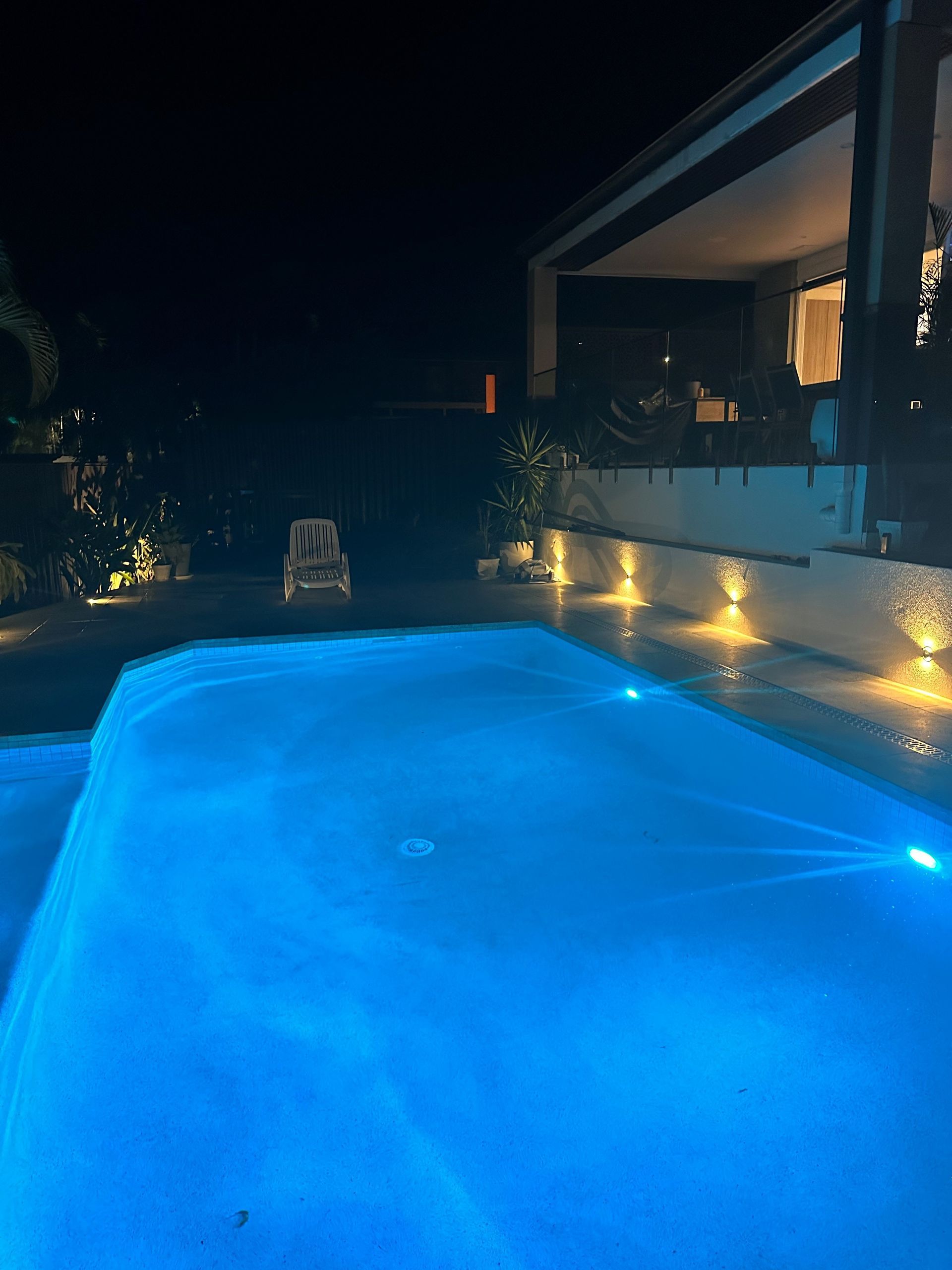 A nighttime view of a pool lit with blue lights. The pool reflects the glow with a house in the background.