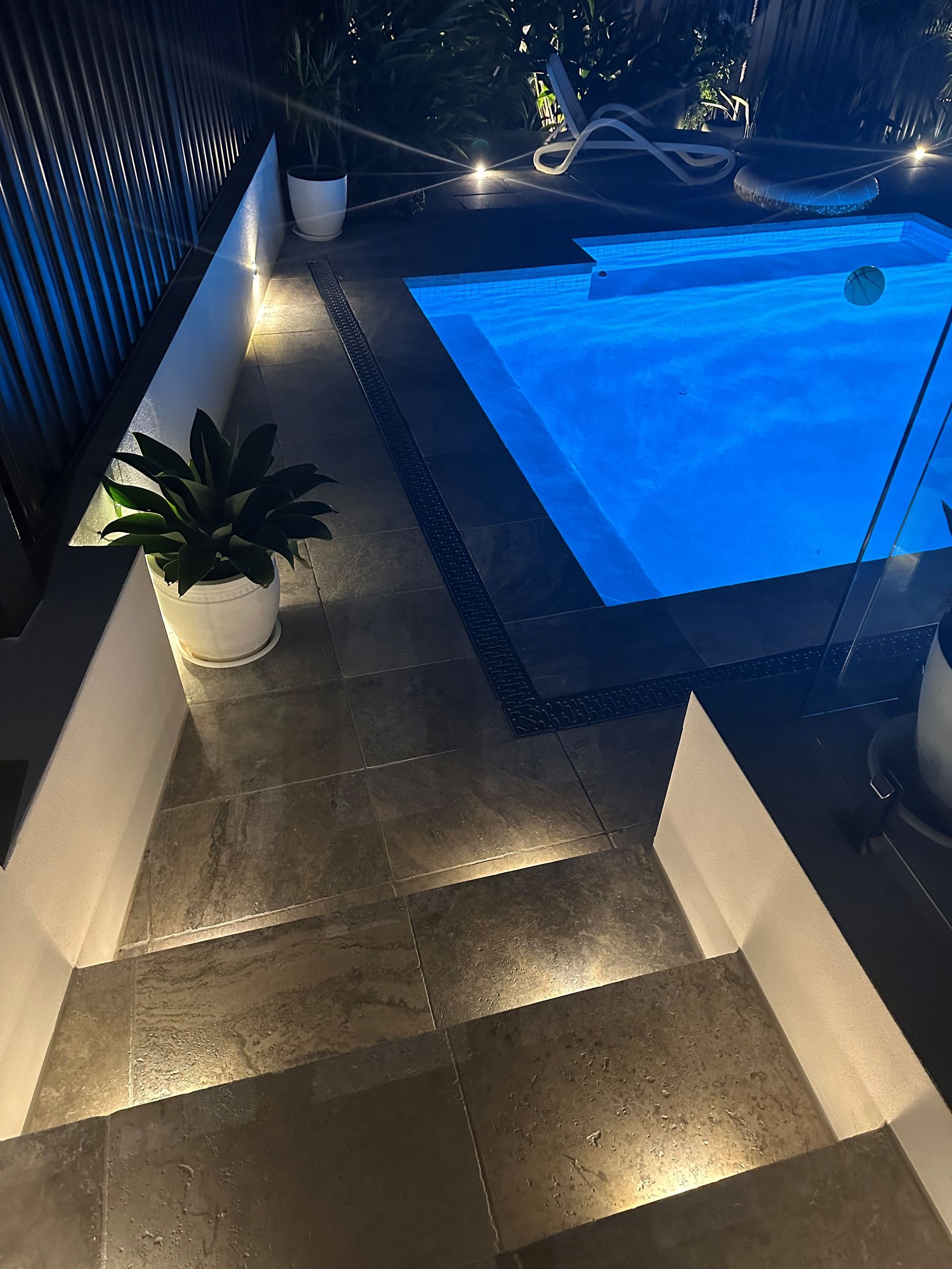Dark patio with steps leading to a pool, lit by small ground lights. Plants in white pots.