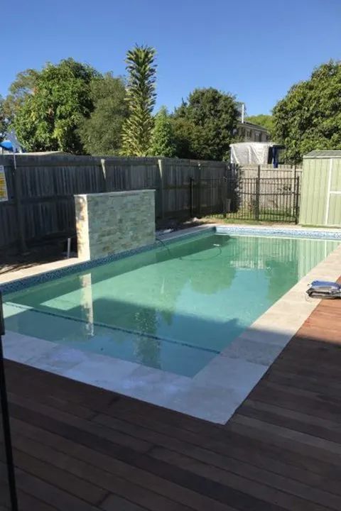 Pool with stone water feature and wooden deck, surrounded by a fence and trees on a sunny day.