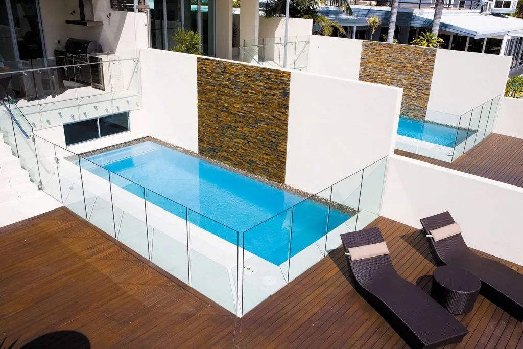 Rooftop pools with glass railings and lounge chairs on wooden decking.