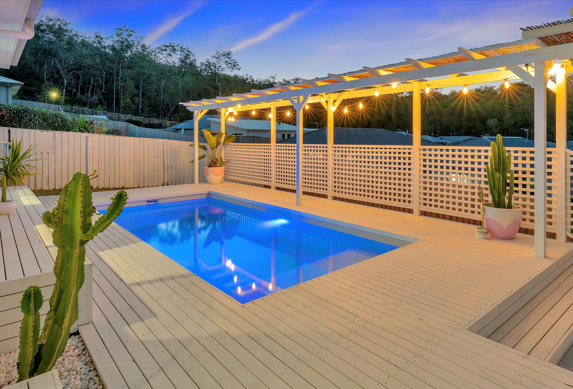 Backyard pool at dusk with white deck, fence, and pergola with string lights.