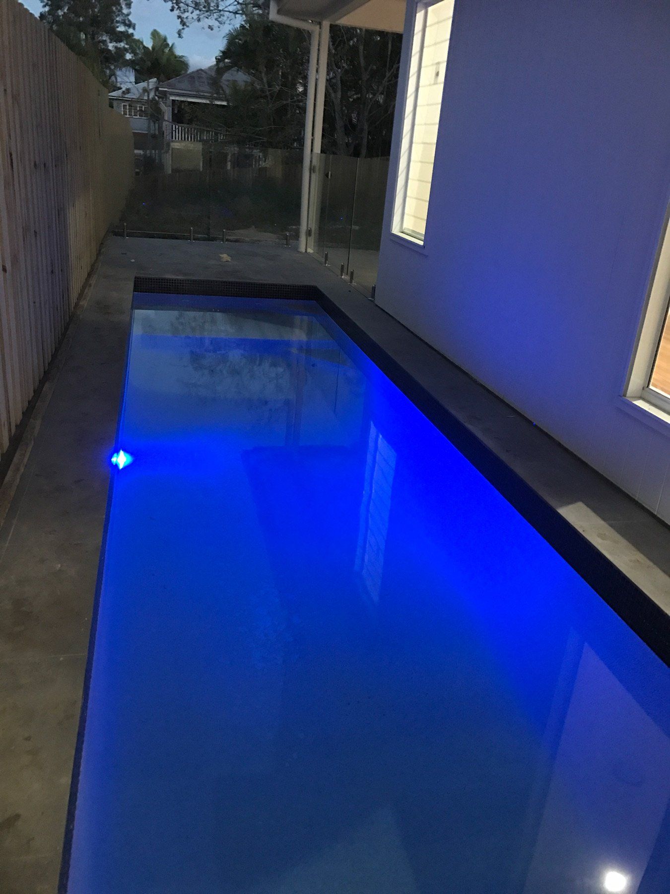 Narrow rectangular pool with blue water, between a white building and a wooden fence.
