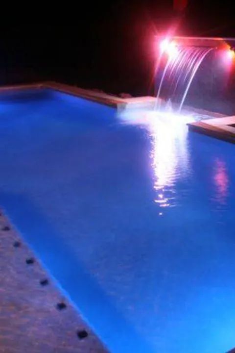 Blue illuminated swimming pool with waterfall feature at night.