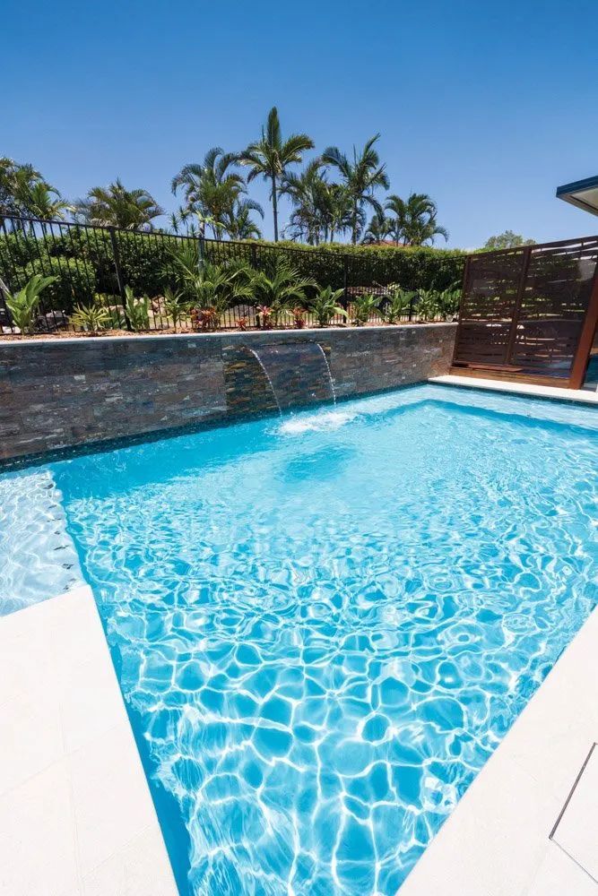 Swimming pool with blue water, stone wall waterfall, and palm trees under a clear sky.
