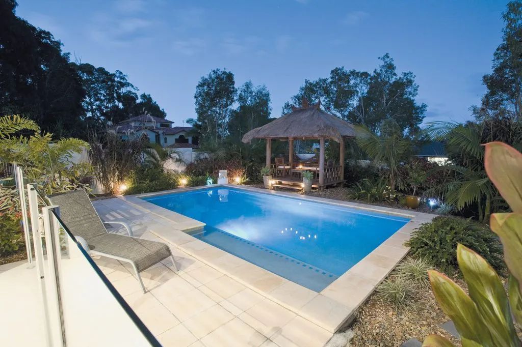 Pool area at dusk with a blue pool, lounge chair, gazebo, and surrounding greenery.