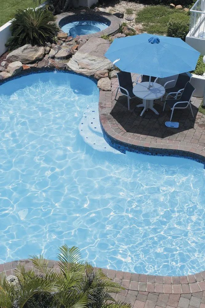 Pool with blue water, hot tub, patio with table, and umbrella, surrounded by brick and landscaping.