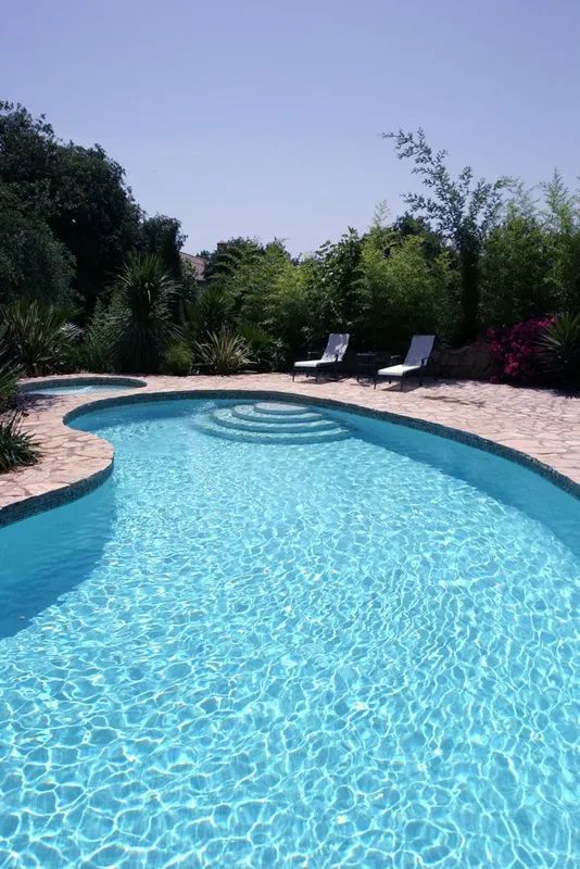 Blue pool with steps, chaise lounges, and surrounding greenery under a bright blue sky.