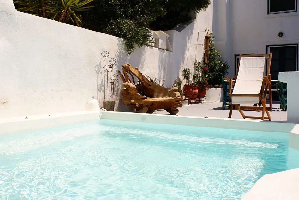 Poolside scene: white walls, clear pool, wooden lounge chair, driftwood, and greenery.