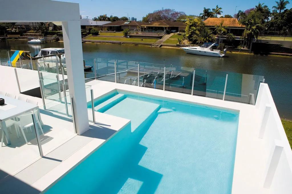 Poolside view with turquoise water, overlooking a waterway.  White deck, glass railings, boats, and suburban homes.