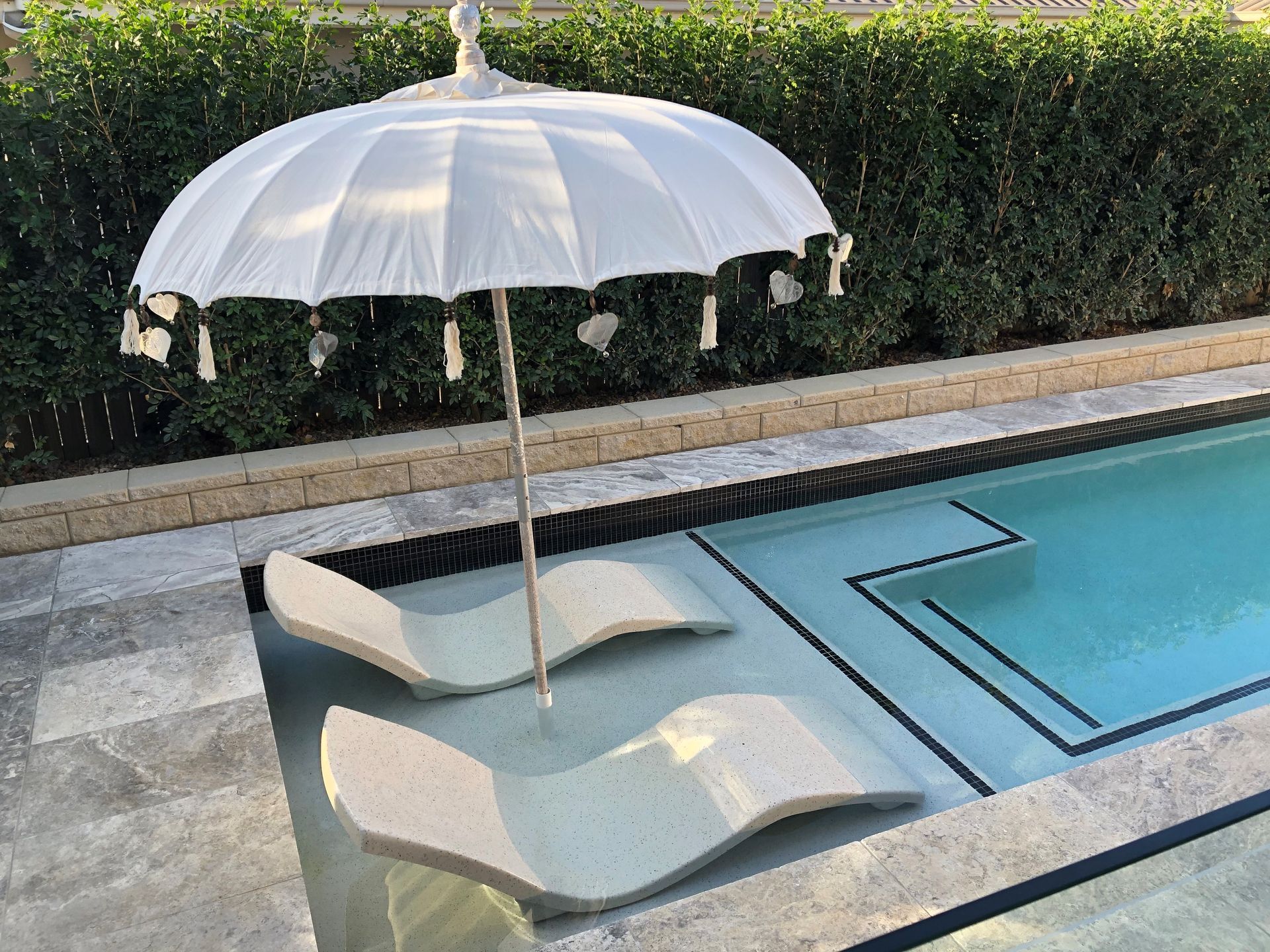 Pool with submerged lounge chairs, white umbrella, and green hedge.