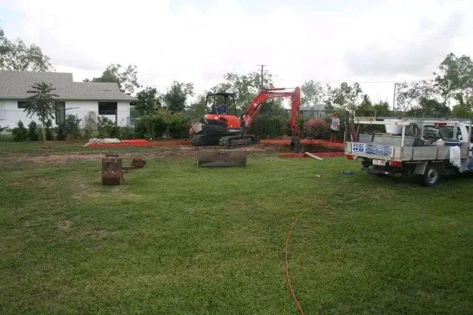 Construction site with excavator digging in a grassy area, a truck, and a house in the background.