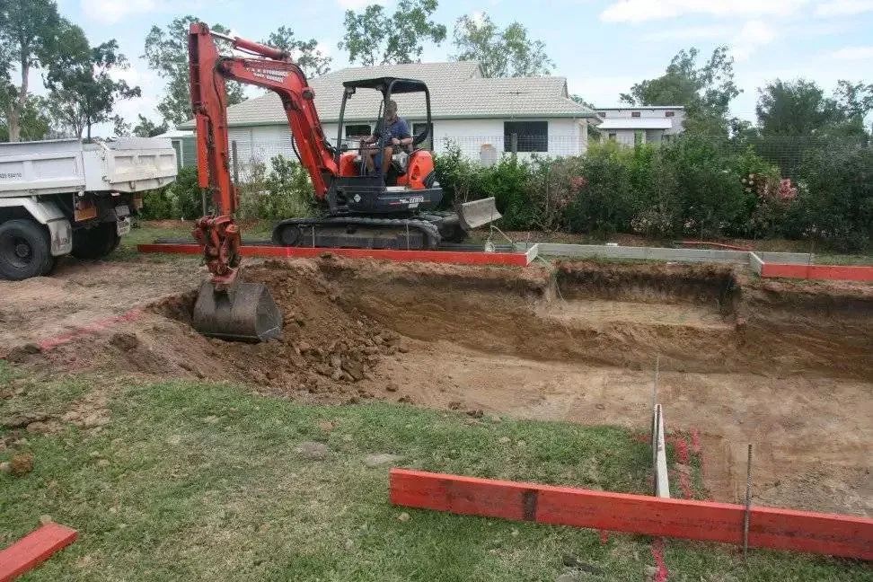 Mini-excavator digging a trench in a yard, with a dump truck nearby and a house in the background.