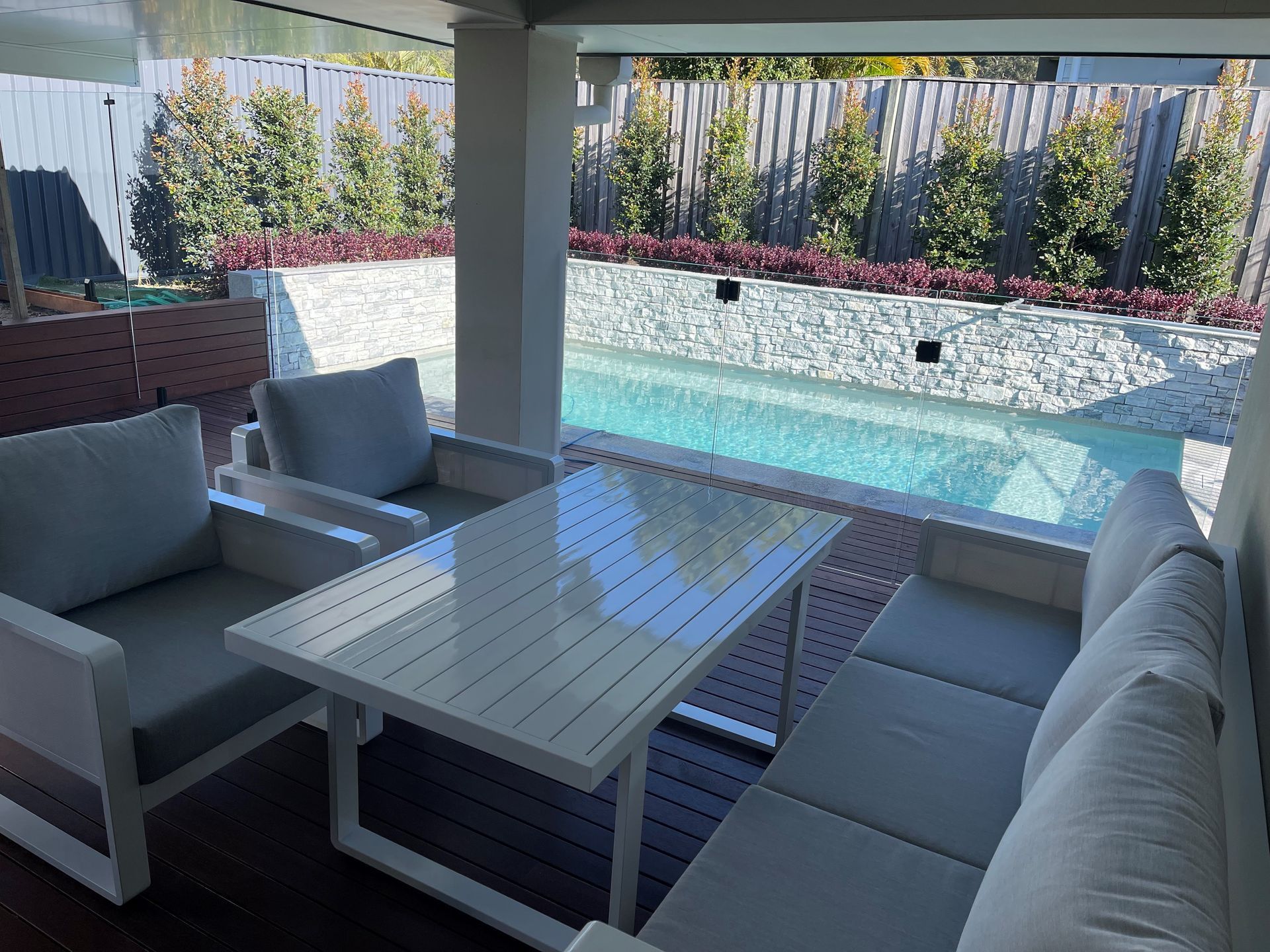 Outdoor seating area with table, cushions, and pool. White furniture, sunny setting.