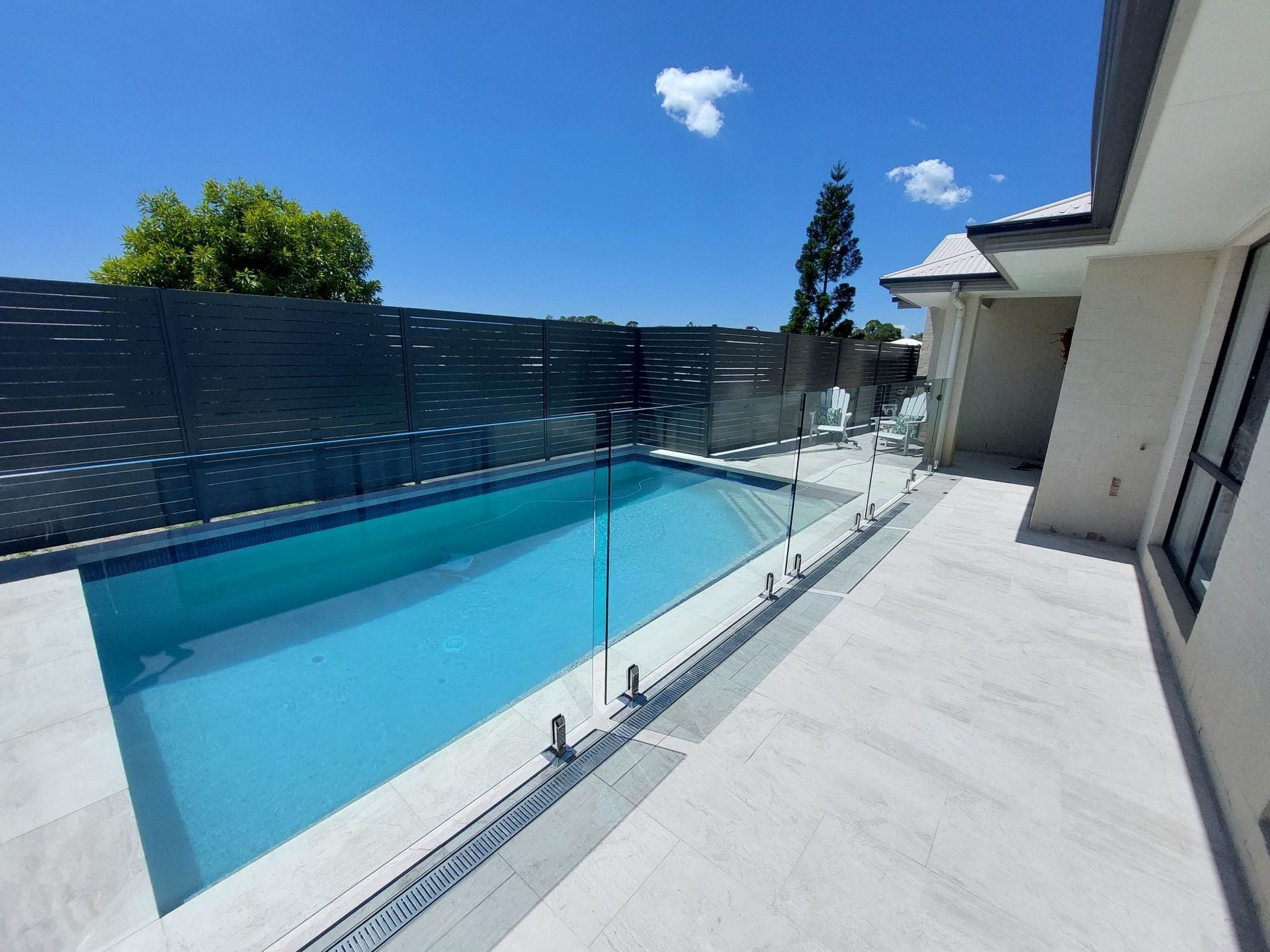 Pool with glass fencing, grey patio, and blue sky.