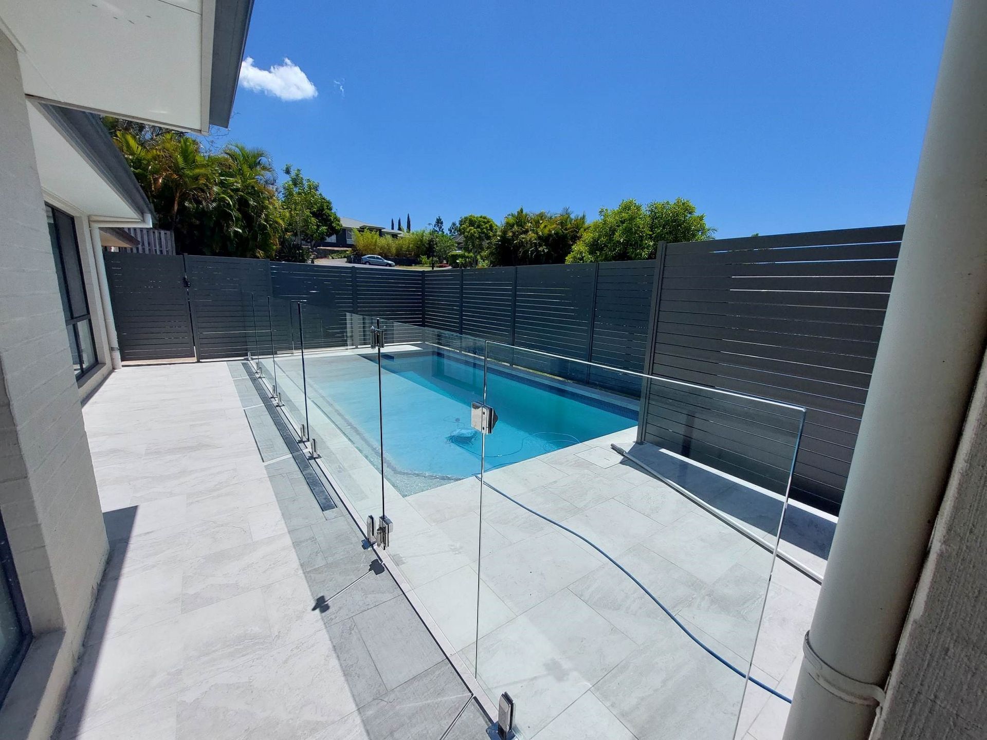 A backyard pool with glass fencing, grey patio, and a dark grey fence against a blue sky.