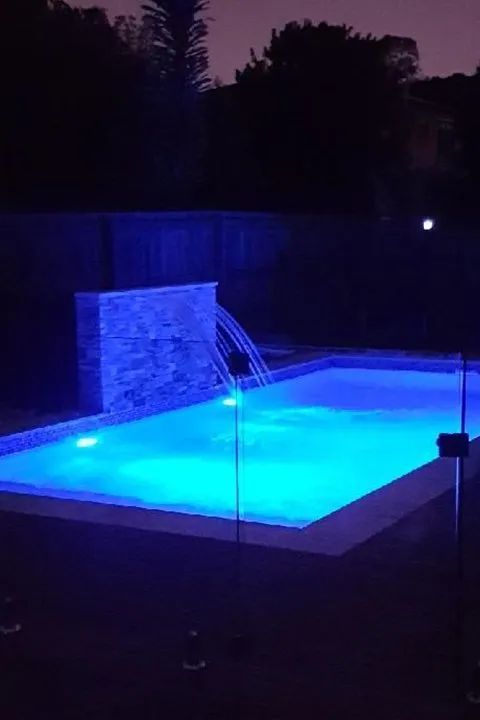A nighttime view of a rectangular pool illuminated with blue light, featuring a waterfall feature and a dark background.