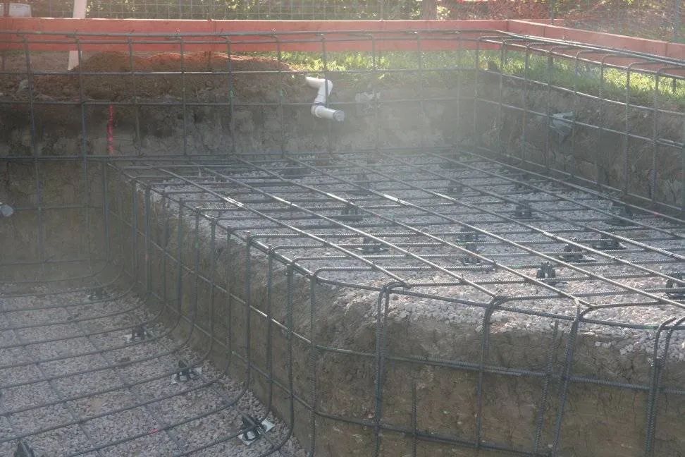 A pool foundation under construction, showing rebar framework, ground, and retaining walls.
