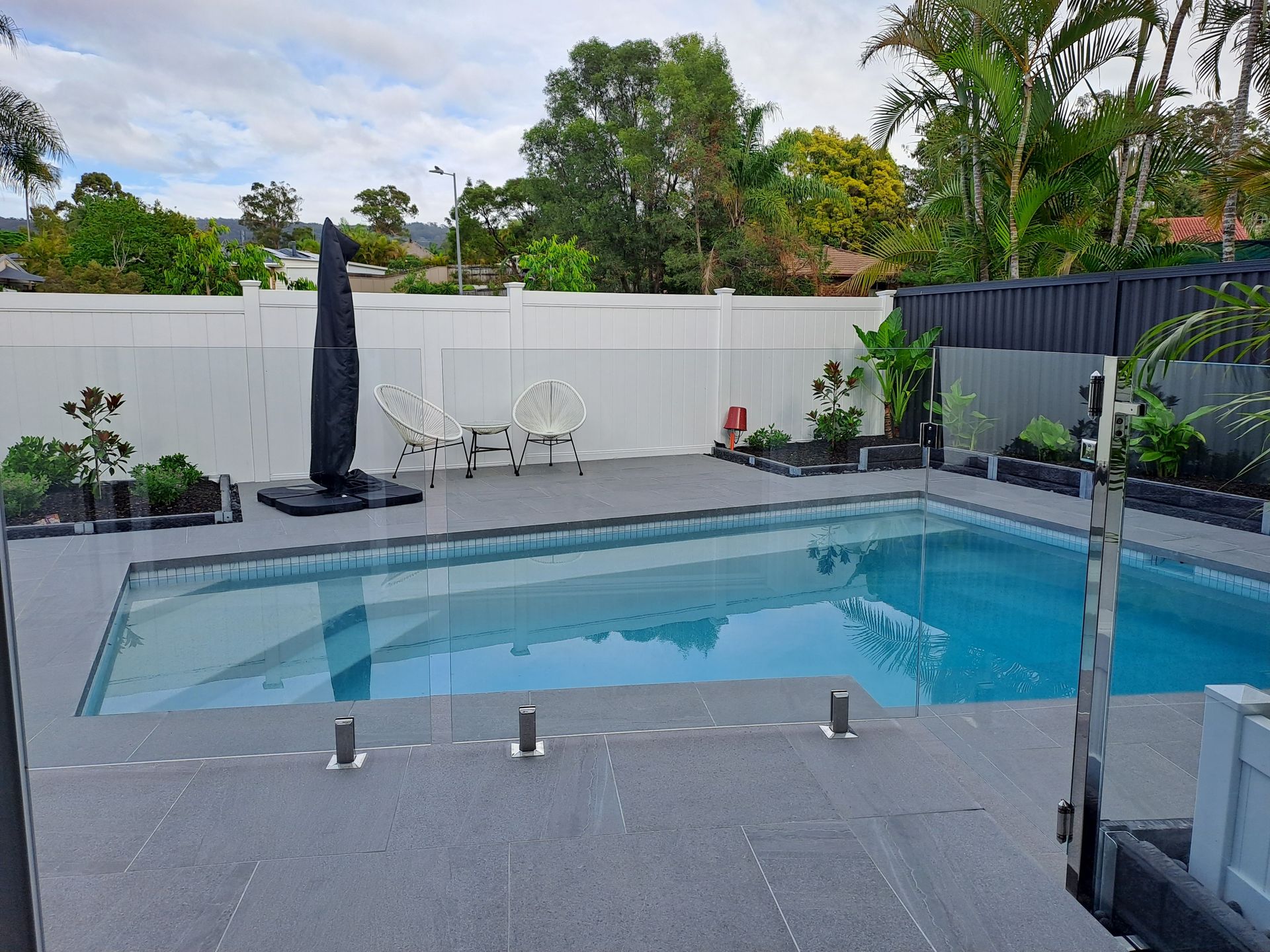 A backyard pool with gray tiling and a white fence. Includes umbrella, chairs, and landscaping.