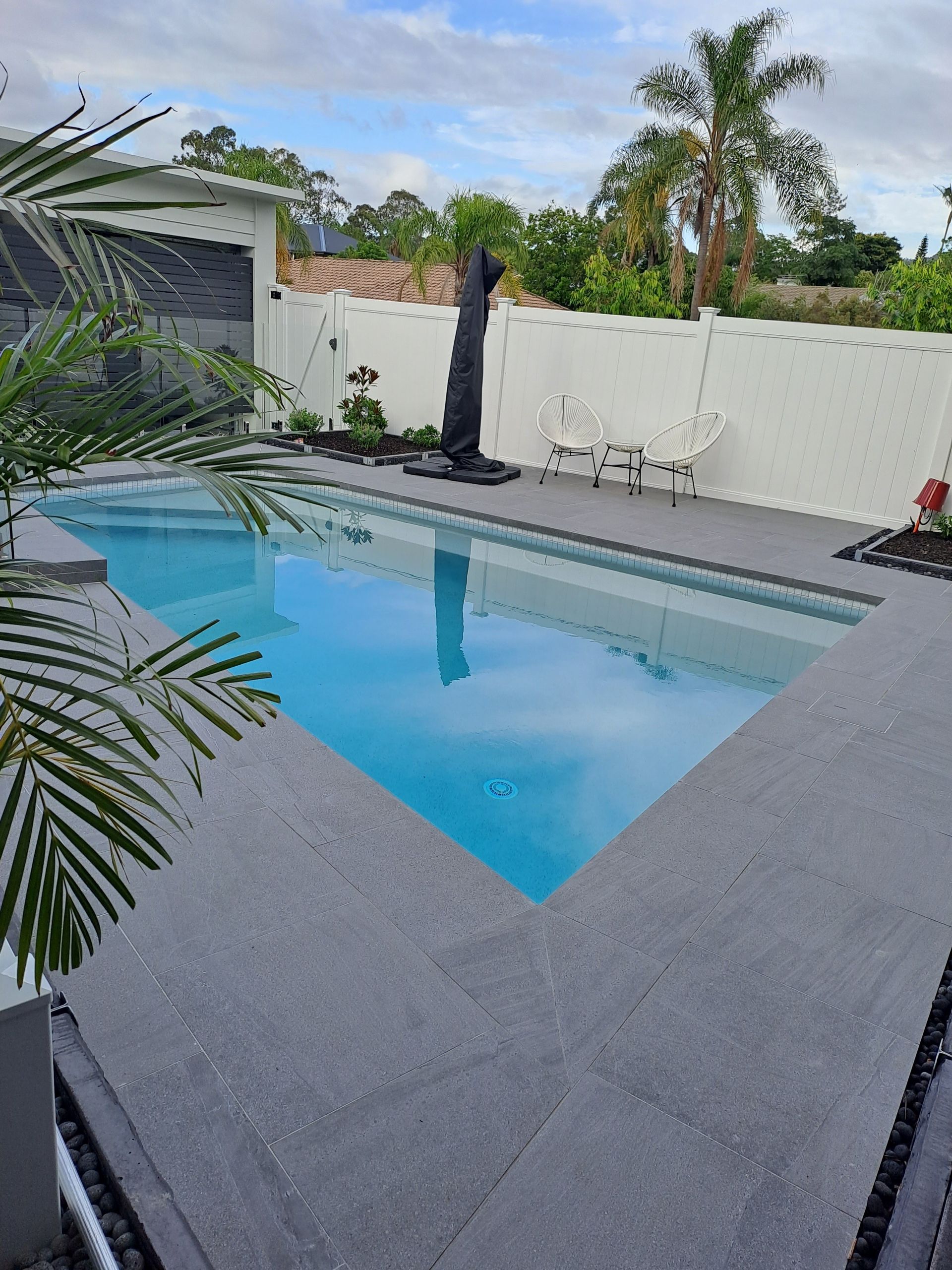 Pool with blue water, gray concrete deck, white fence, and palm trees in the background.