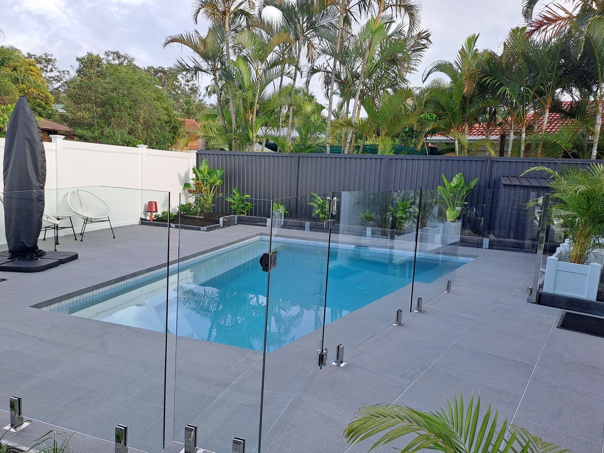 Backyard pool surrounded by a gray deck, glass fencing, and lush green plants. Black fence and umbrella.