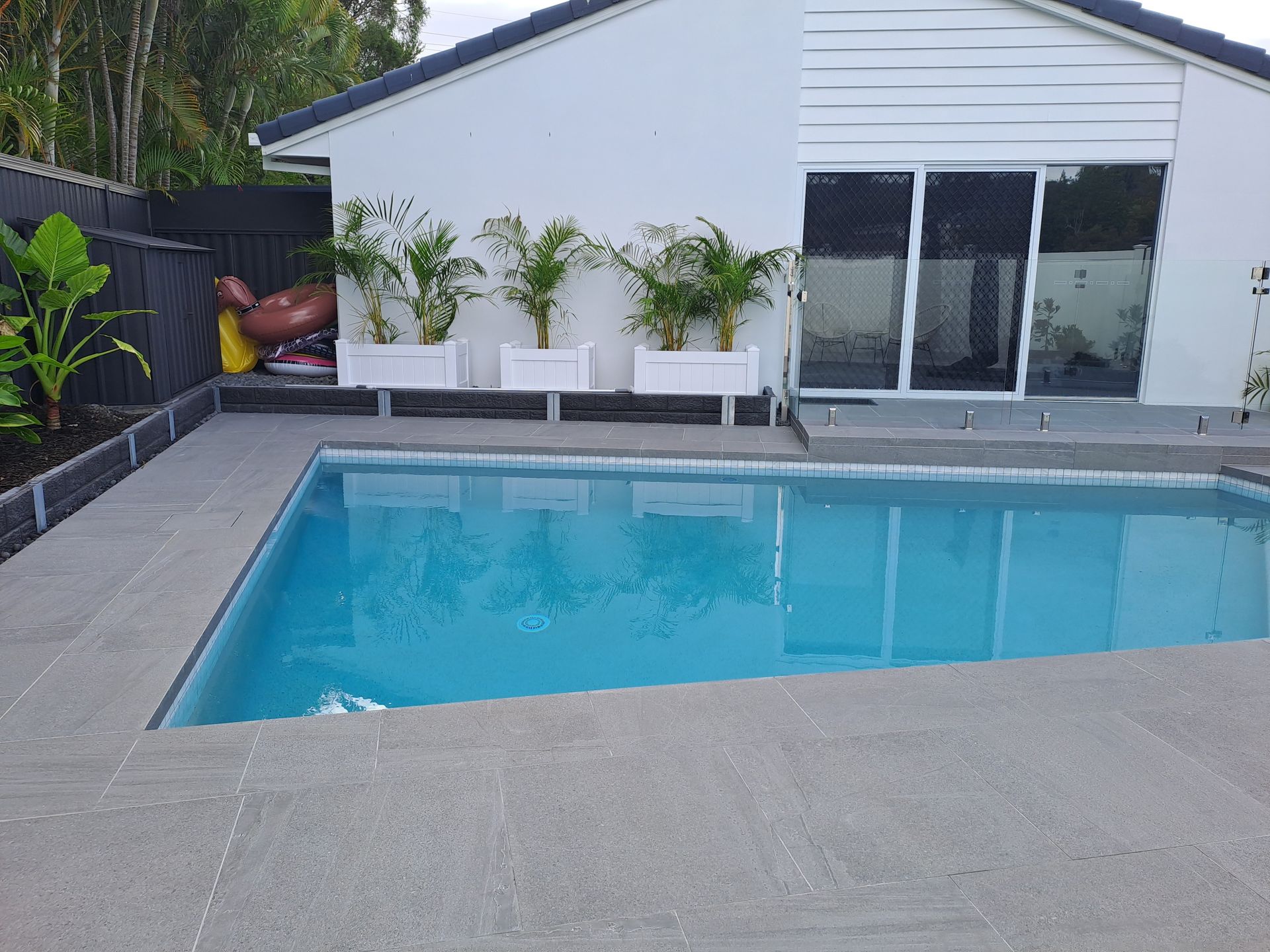 A backyard pool with gray concrete surround; white house and planters with green plants in the background.