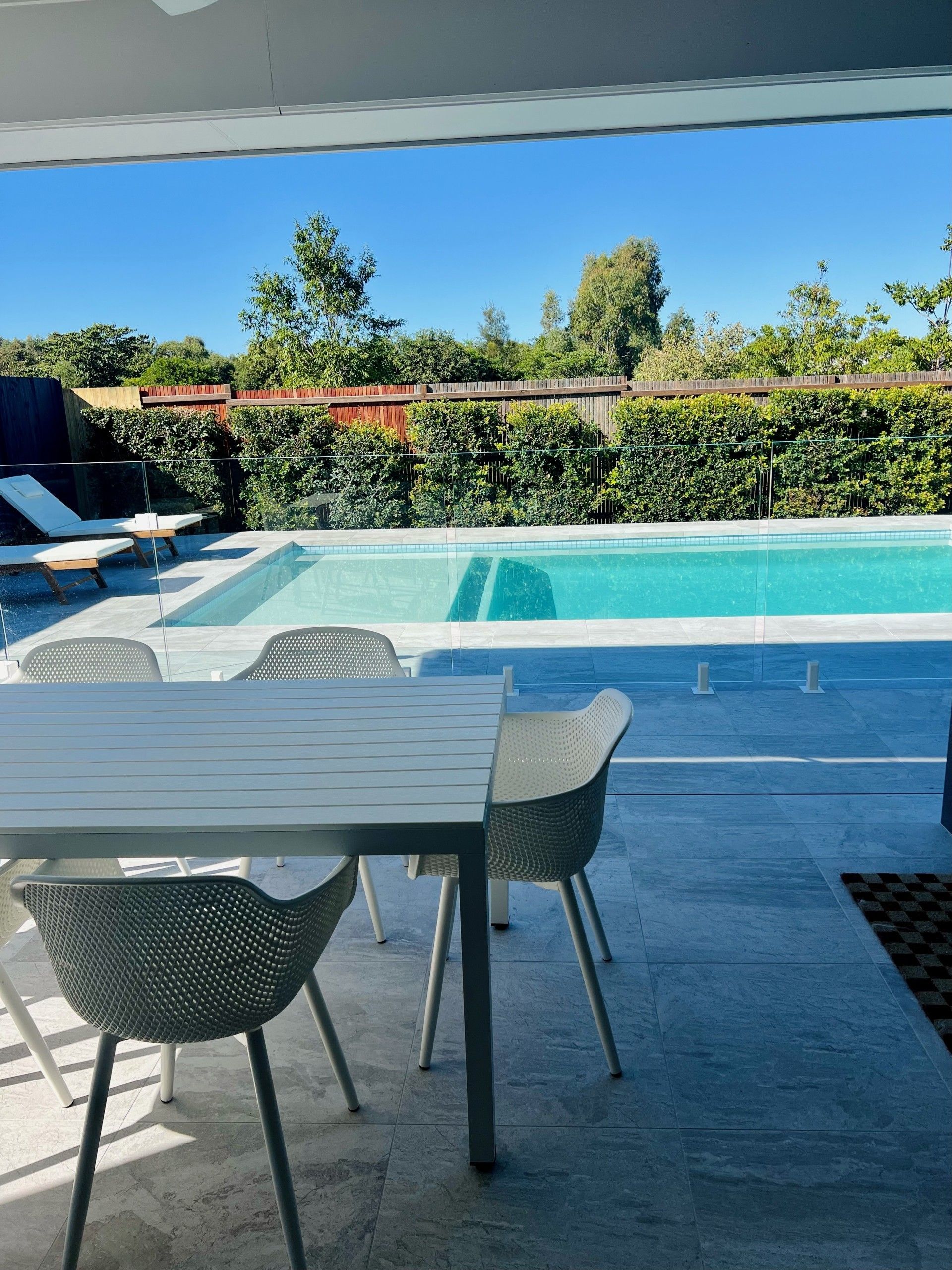 Poolside patio with table and chairs overlooking a rectangular pool and lush green hedge under a blue sky.