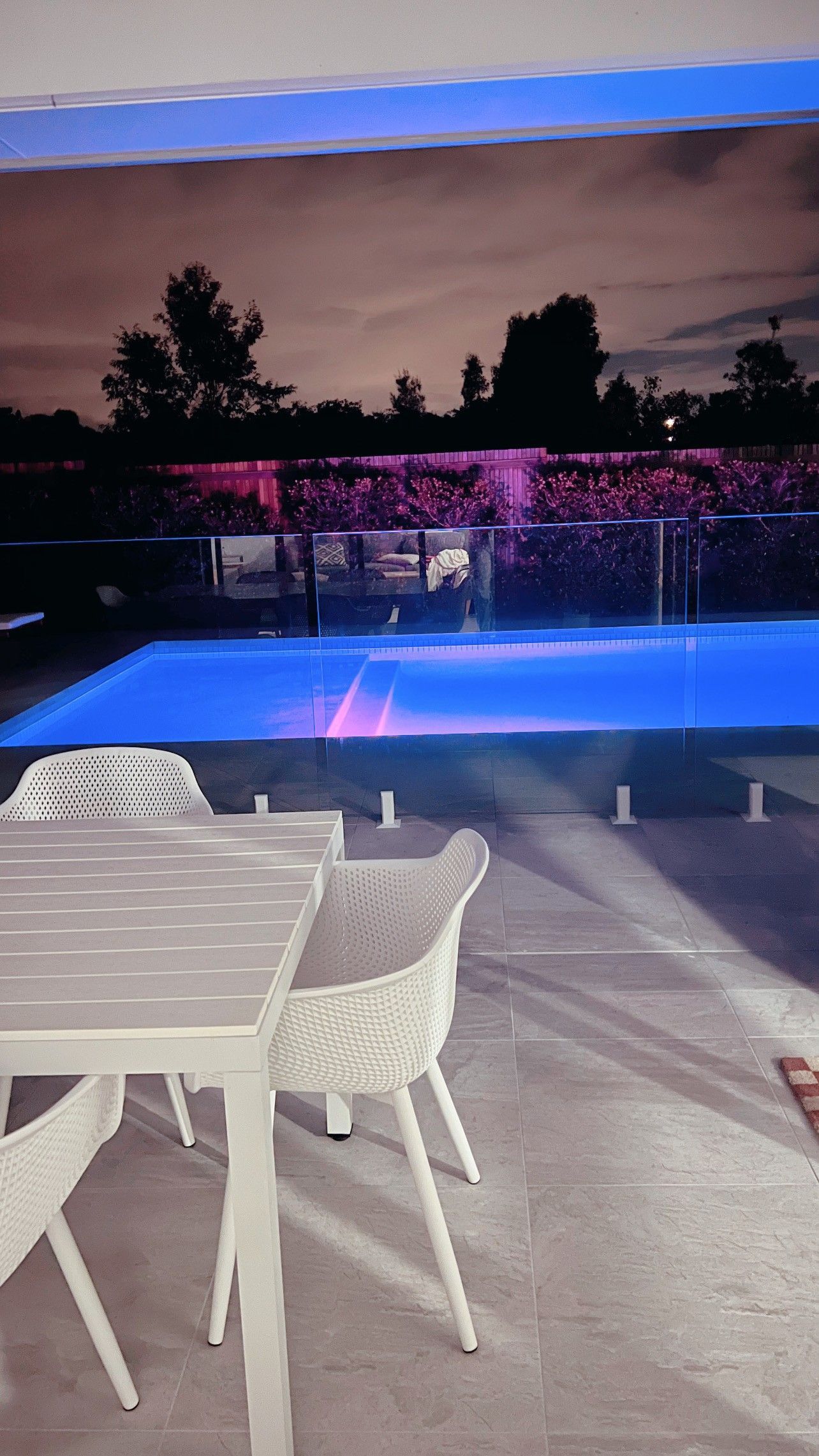 A nighttime poolside scene with a table and chairs, with a lit up pool and trees in the background.