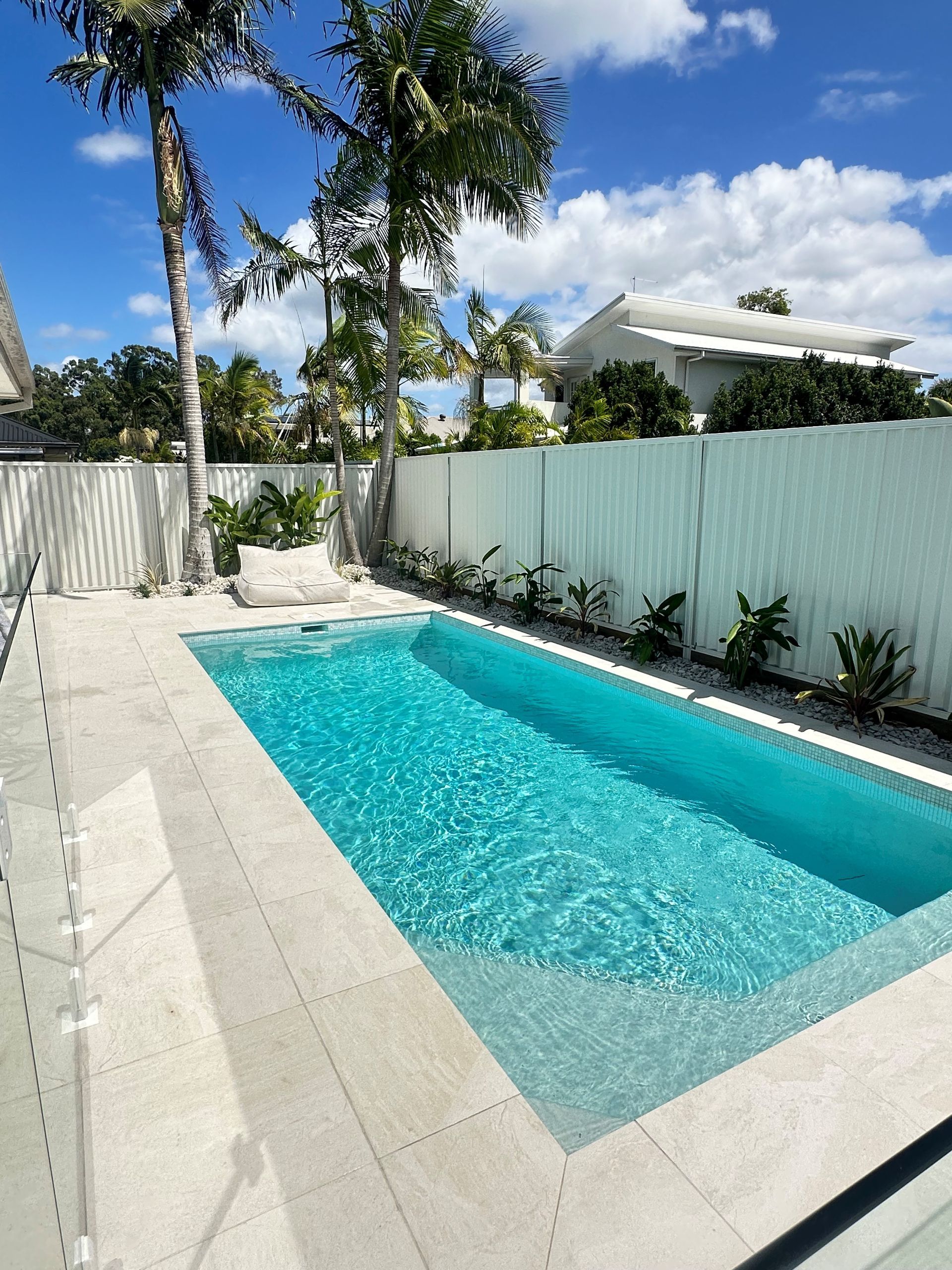 A rectangular pool with turquoise water, framed by light-colored stone, palm trees, and a white fence under a blue sky.