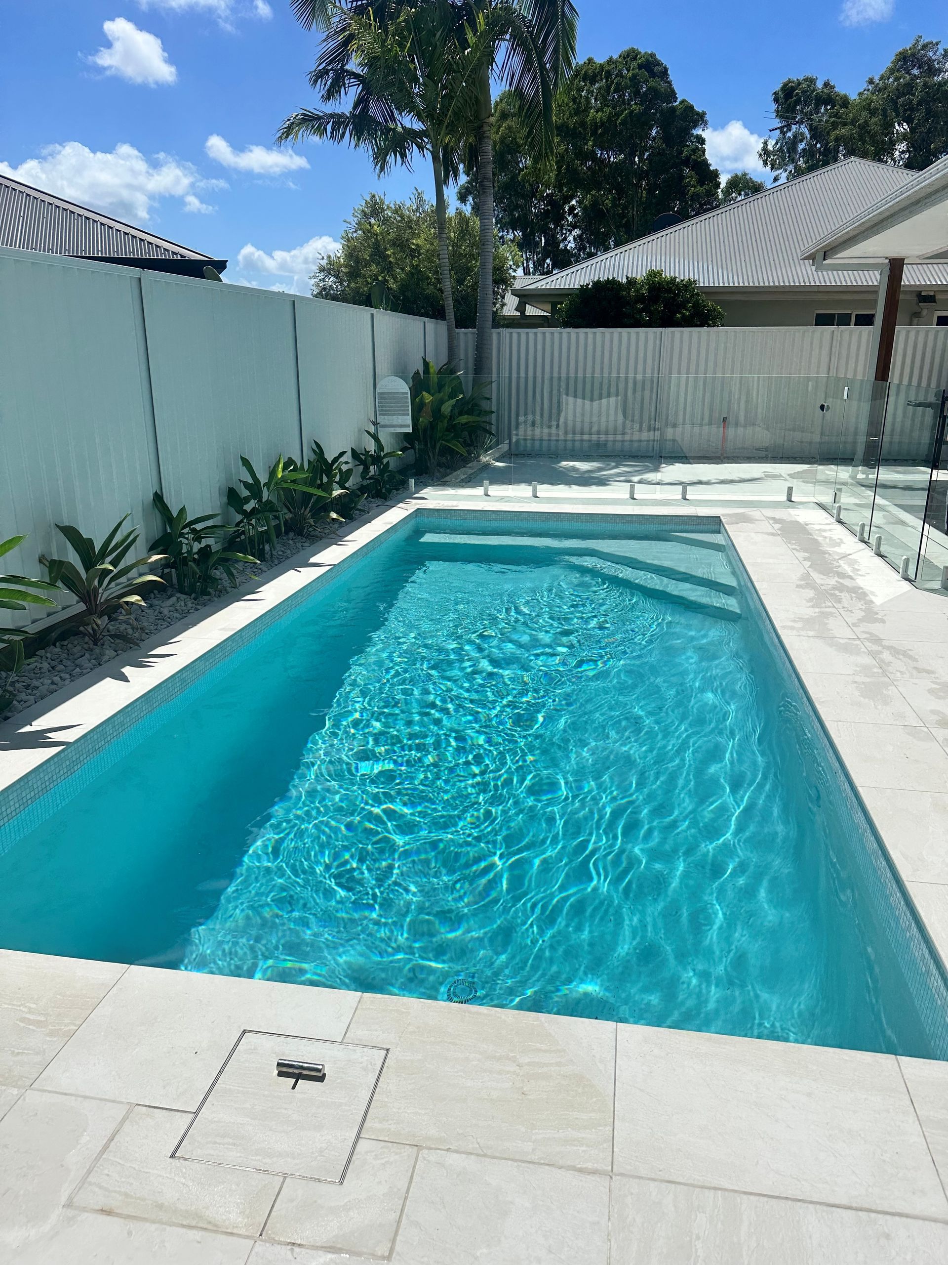 Rectangular turquoise pool in a backyard with white fence and stone patio.
