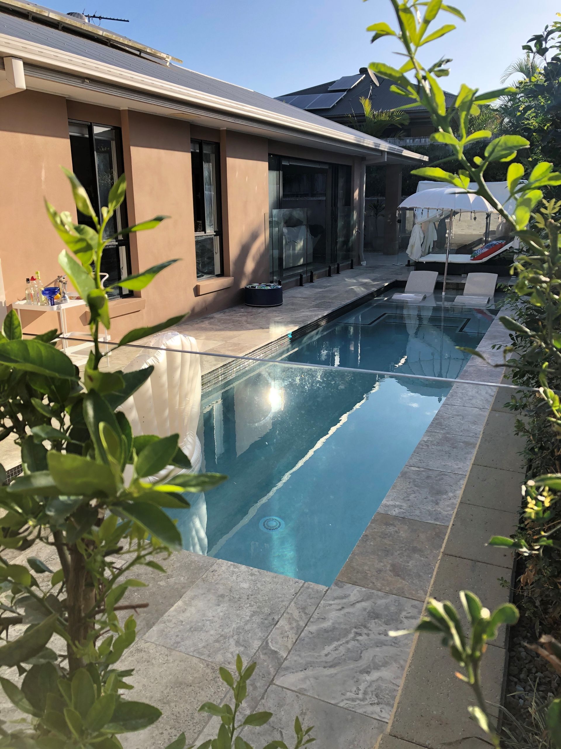 Pool next to a house; blue water, tan tile, lush green plants. Sunny day.