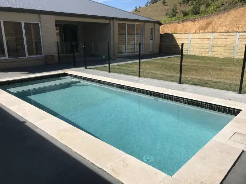 Rectangular swimming pool with light blue water, surrounded by gray concrete and a black fence. House in the background.