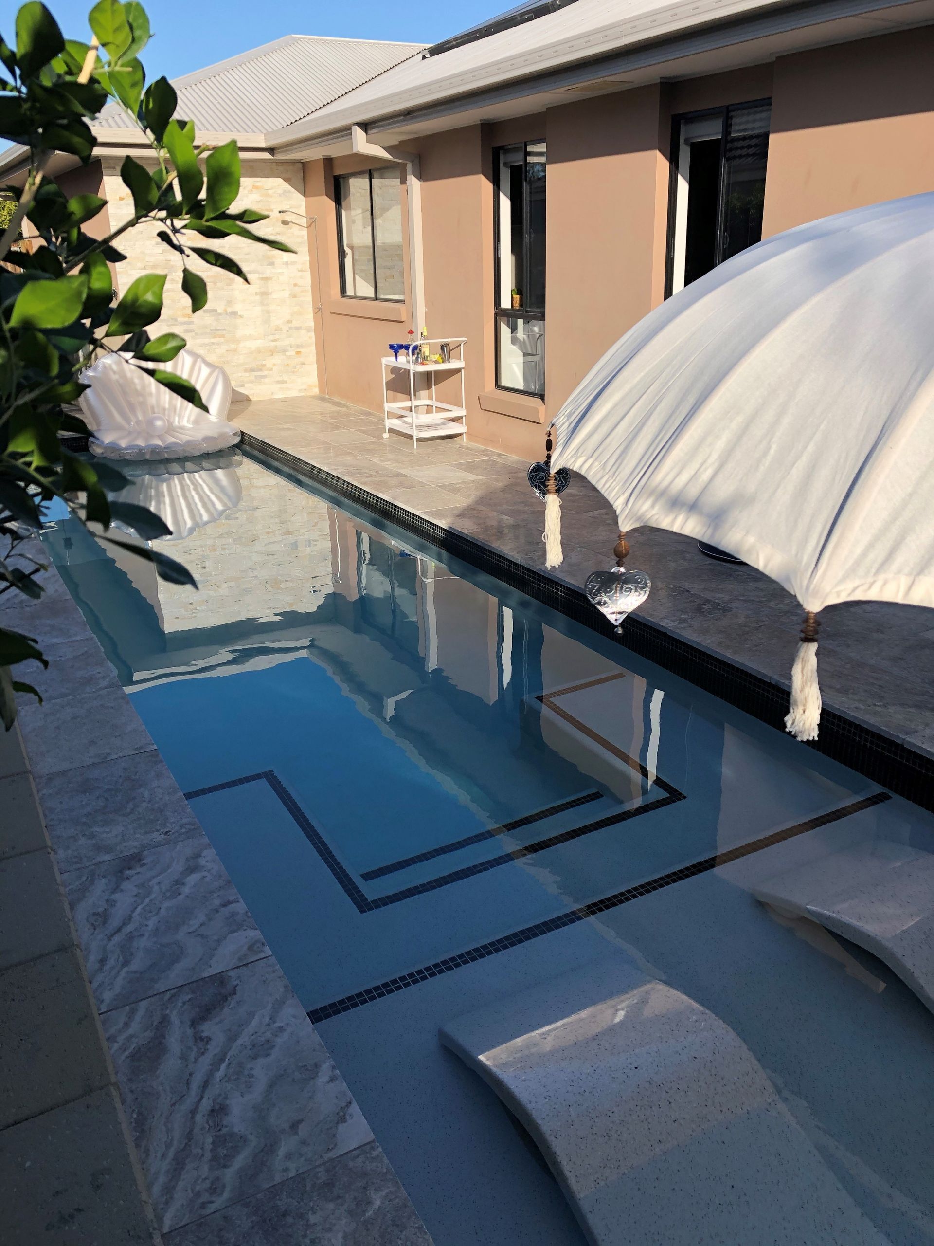 Poolside scene: long rectangular pool, lounge chairs, parasol, and a light brown house in the background.