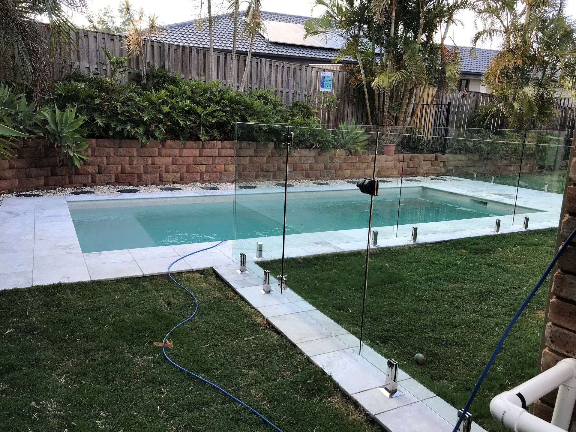 Pool with glass fencing, surrounded by grass, stone pavers, and brick wall.