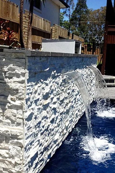 Stone waterfall cascades into a blue swimming pool; sunny outdoor setting.