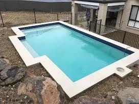 Rectangular in-ground pool with white border. Blue water. Fenced backyard. Adjacent house.
