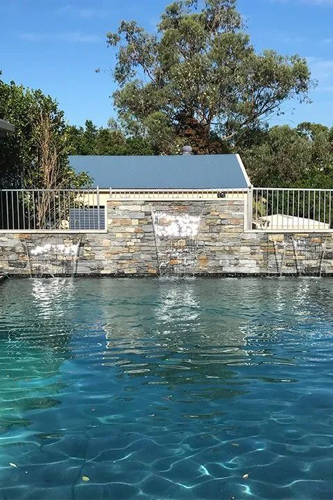 Pool with stone wall, water cascading down, blue sky, building and trees in the background.