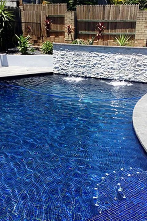 Blue-tiled swimming pool with a stone waterfall feature in a backyard setting.