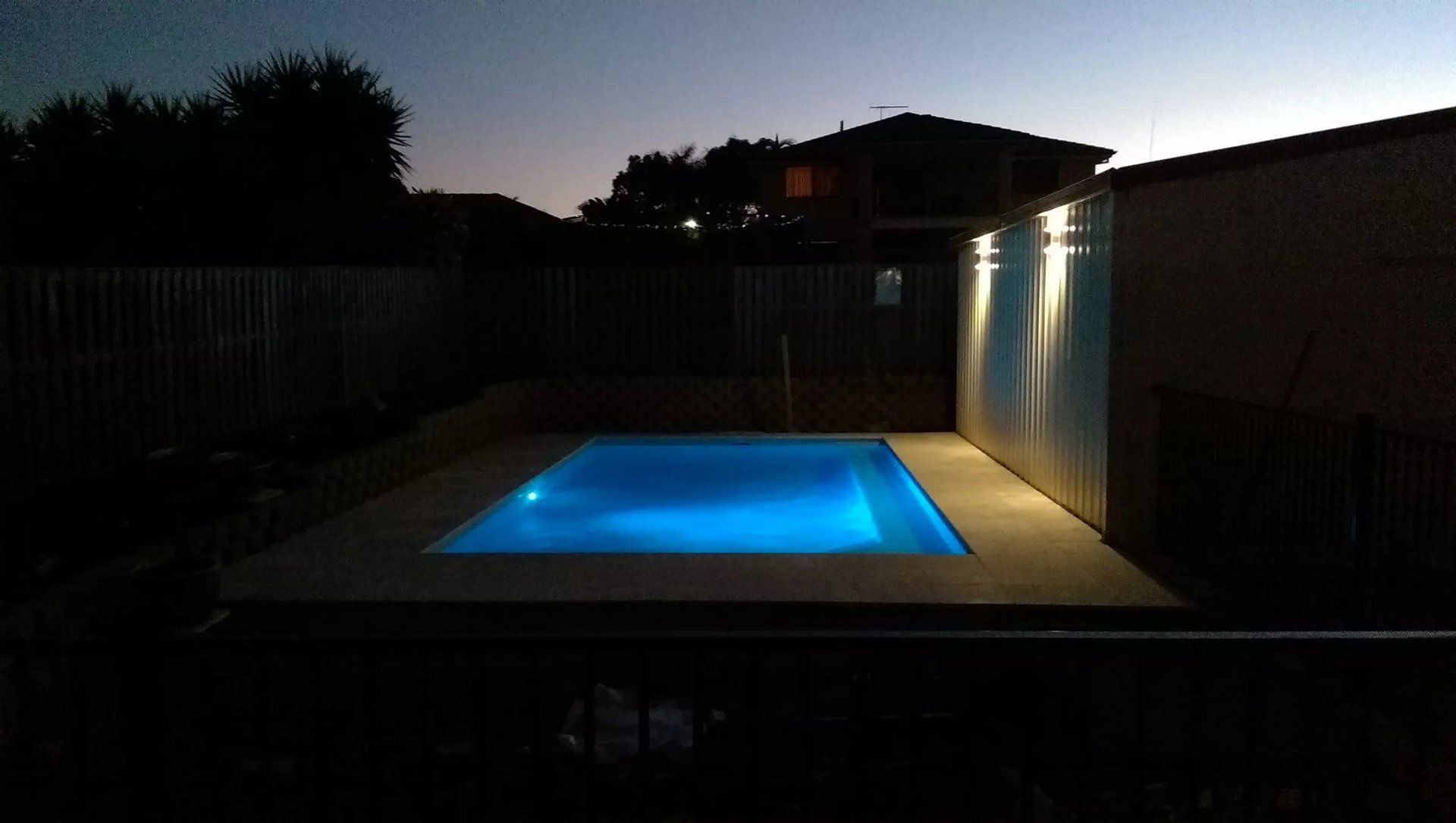 Nighttime view of a backyard pool, illuminated blue, surrounded by a dark fence and building.