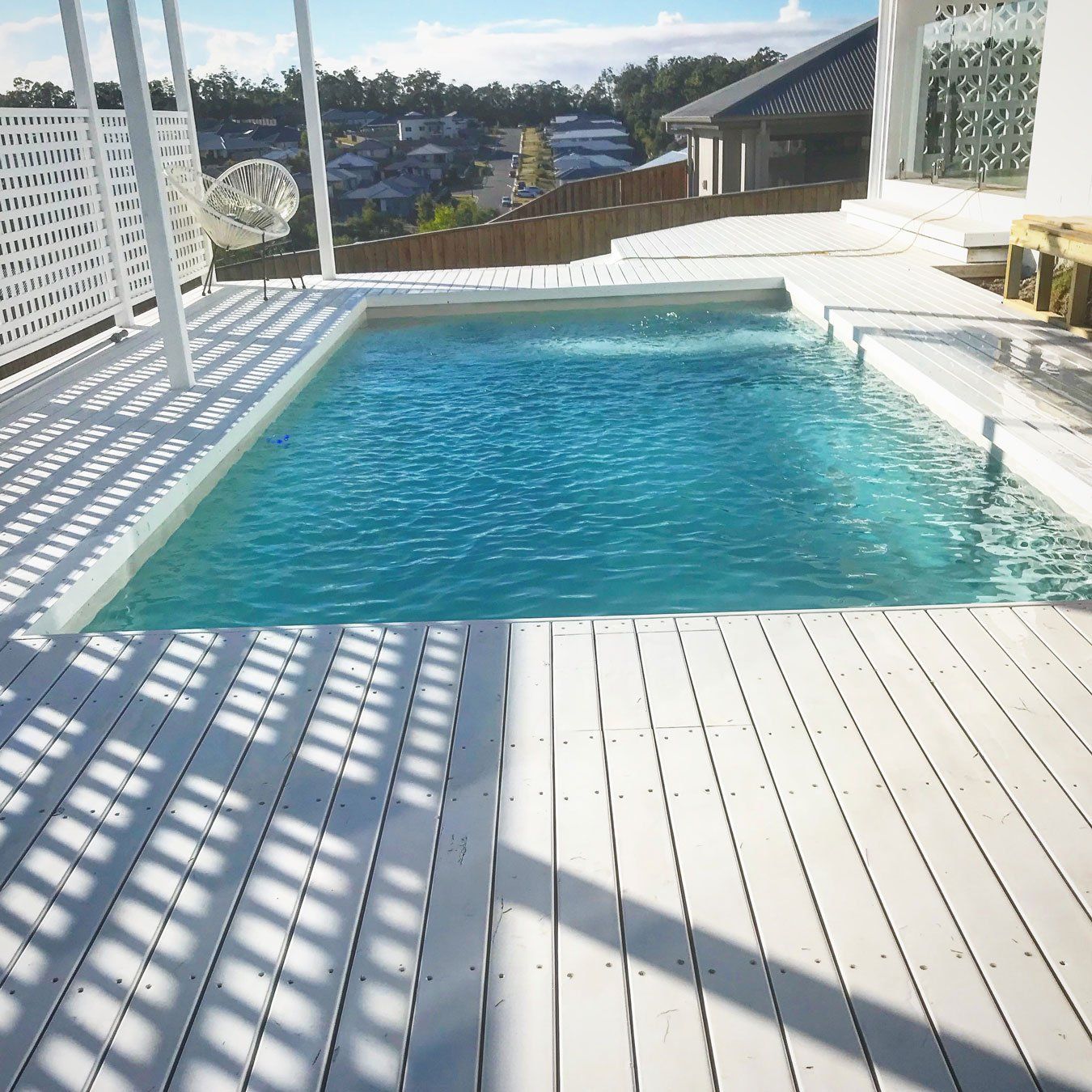White-decked pool with turquoise water, surrounded by white railings, set against a hillside with houses.
