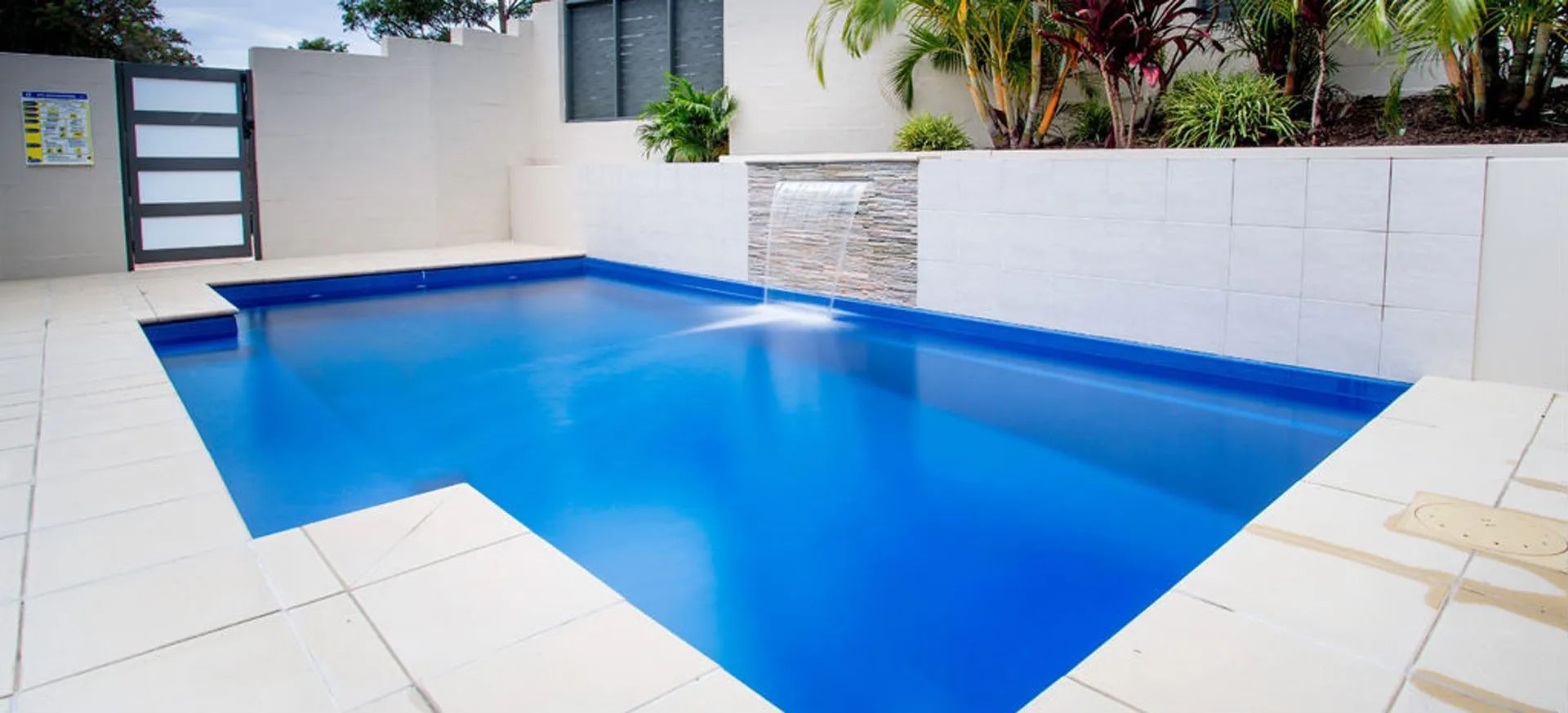 A rectangular blue swimming pool surrounded by white tiles and walls; a waterfall and a gate in the background.