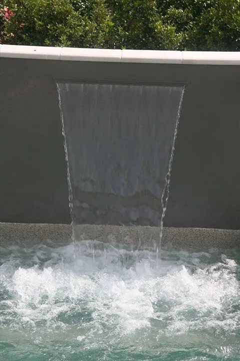 Waterfall cascading into a pool with bubbling water. Gray wall and green foliage in the background.