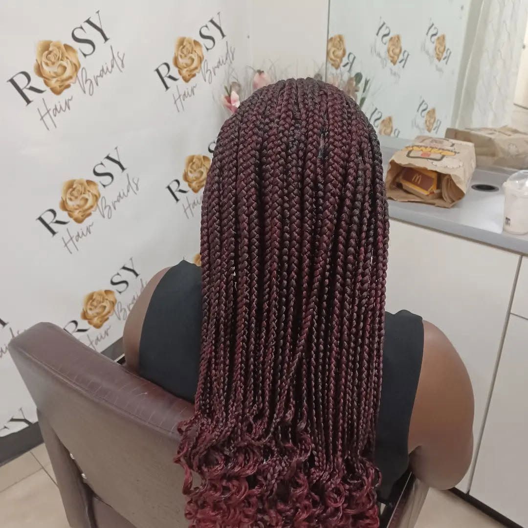 A woman with braids is sitting in a chair in front of a wall that says rosy hair beauty