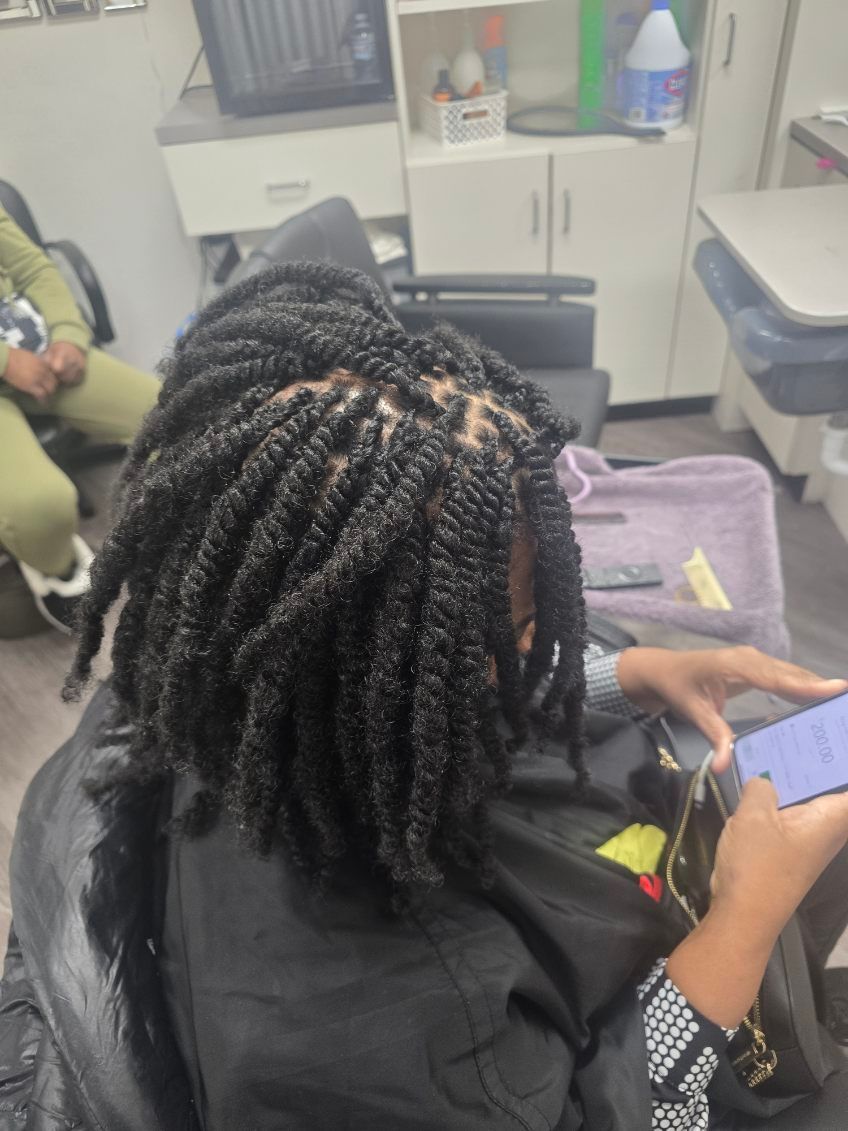 Woman with dark hair styled in twists, sitting in a salon chair, looking at her phone.