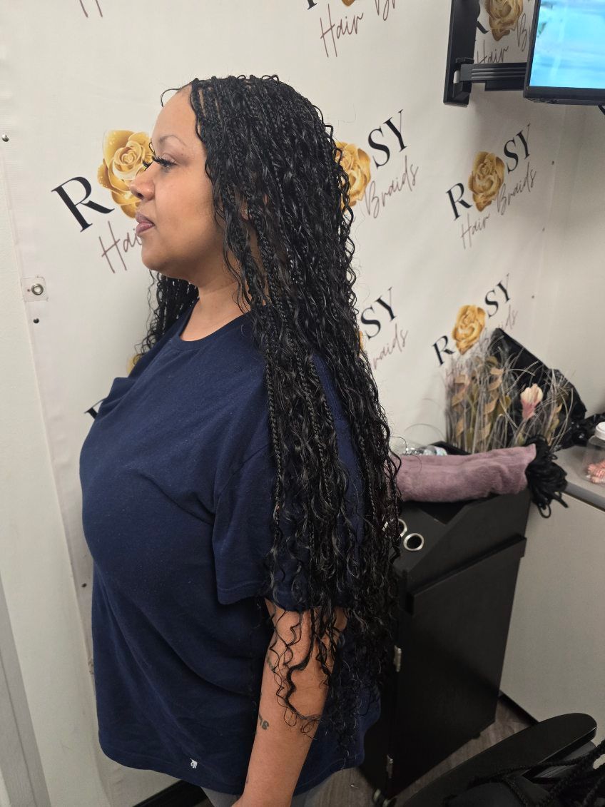 Woman with long, curly black hair extensions, wearing a blue shirt, standing indoors in a beauty salon.