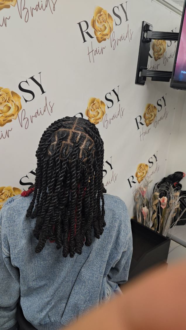 Person with long, dark twists in a salon setting.  The background features a logo with 