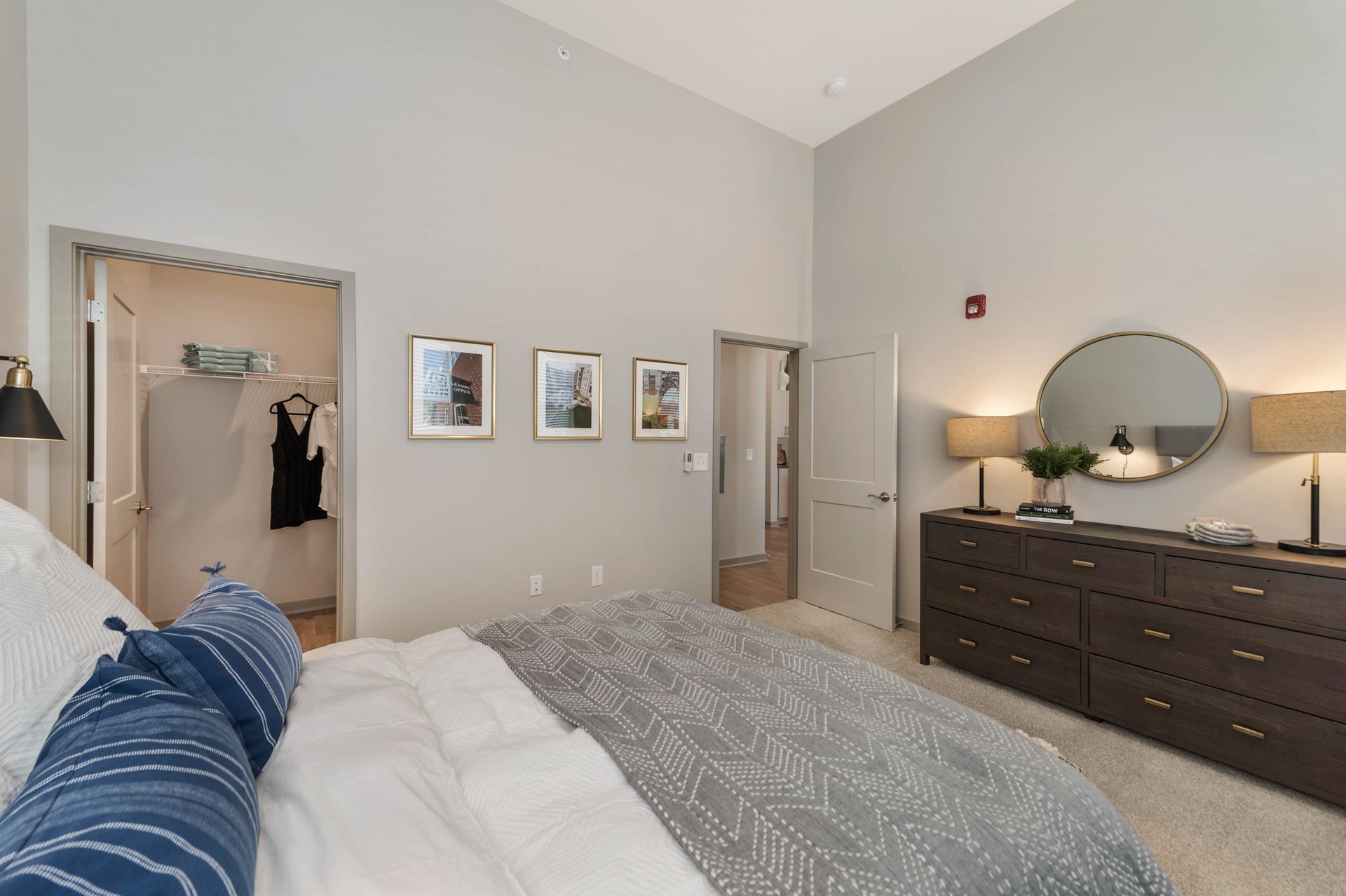 Cozy bedroom featuring a large bed with textured bedding, a dresser with lamps, framed artwork on the wall, and an open closet in the background.