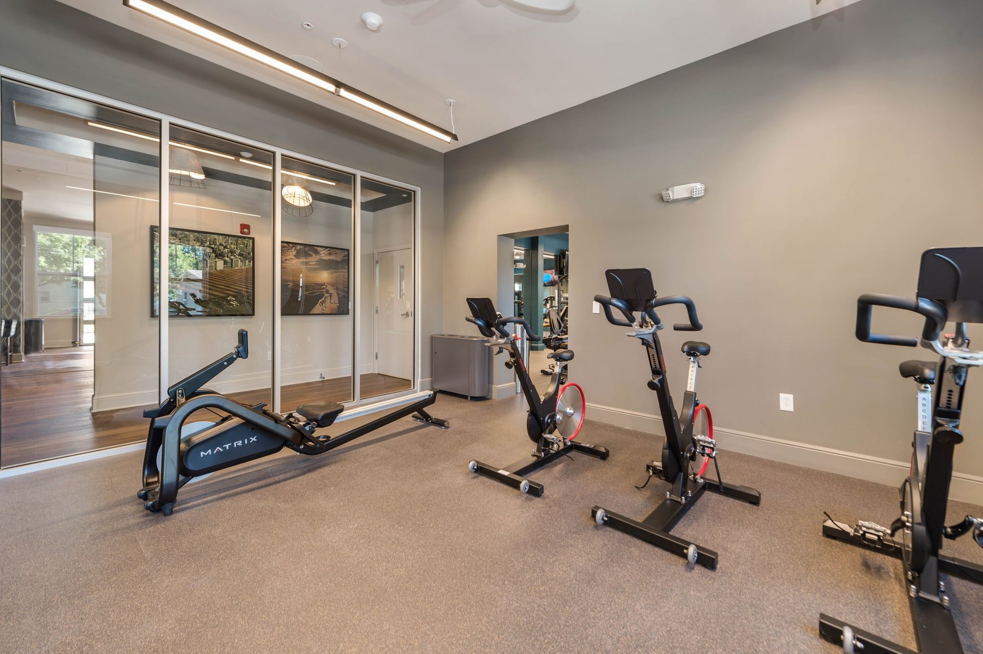 A modern fitness room featuring a rowing machine and several stationary bikes, with glass walls and hardwood flooring visible in the background.