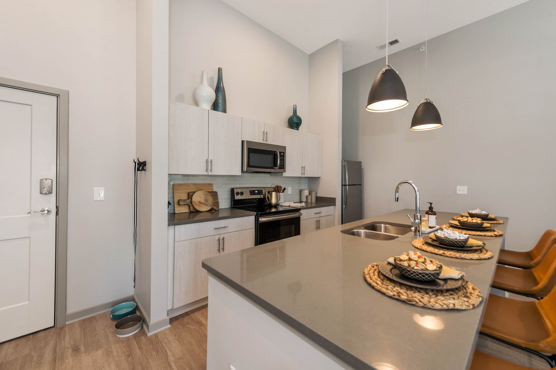 Modern kitchen with light wood cabinets, stainless steel appliances, and a sleek countertop, featuring decorative bowls and pendant lighting above a dining area.
