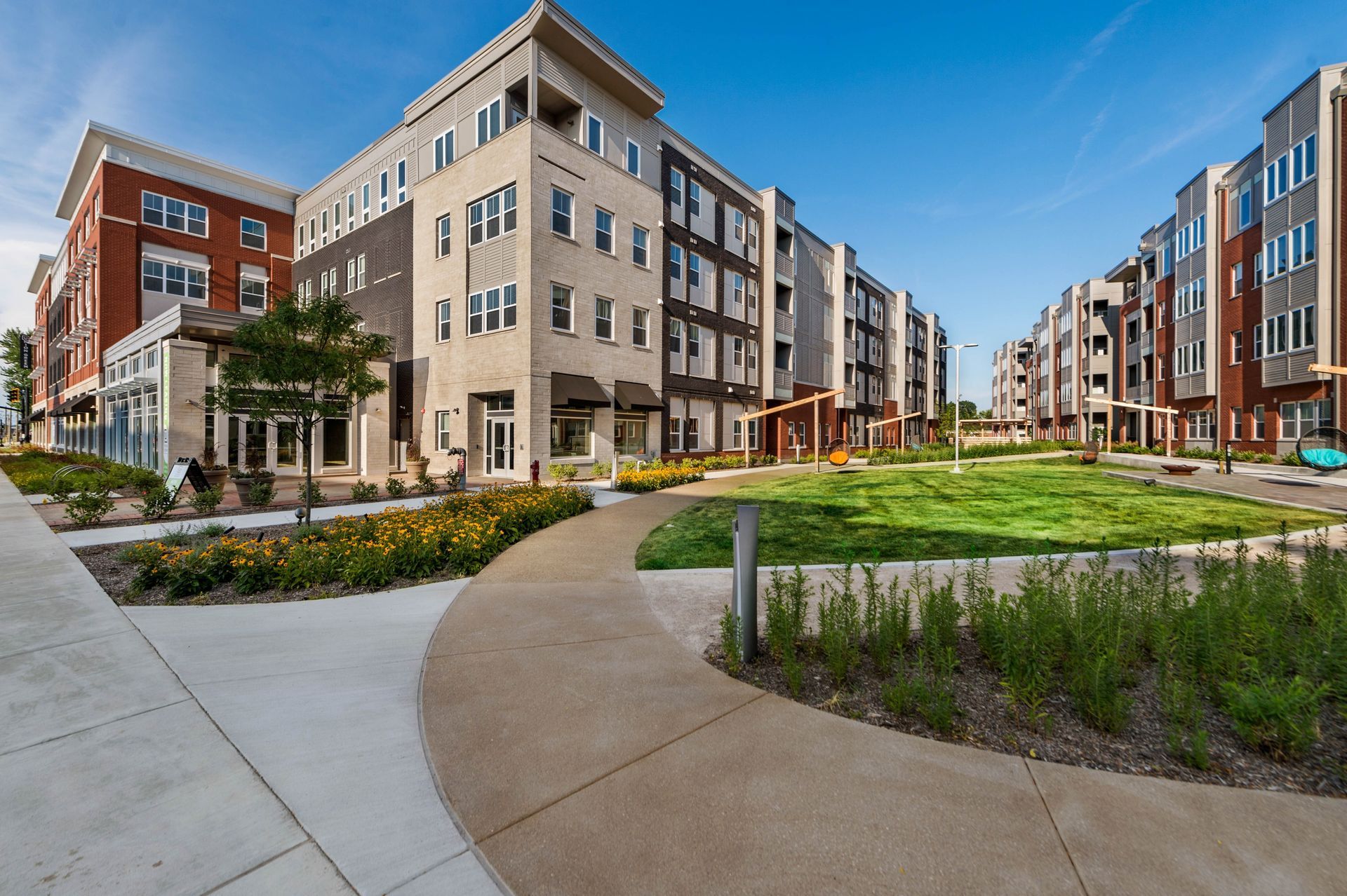 A landscaped residential area featuring two multi-story buildings, a winding pathway, and patches of vibrant flowers and green lawns under a clear blue sky.